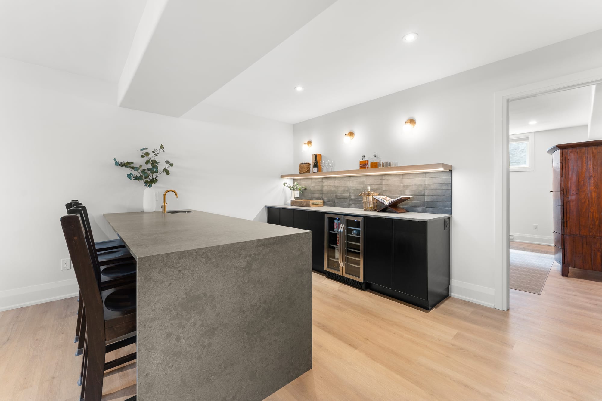 Fully equipped lower-level wet bar with a waterfall stone counter, black cabinetry, dual wine fridges and brass sconces