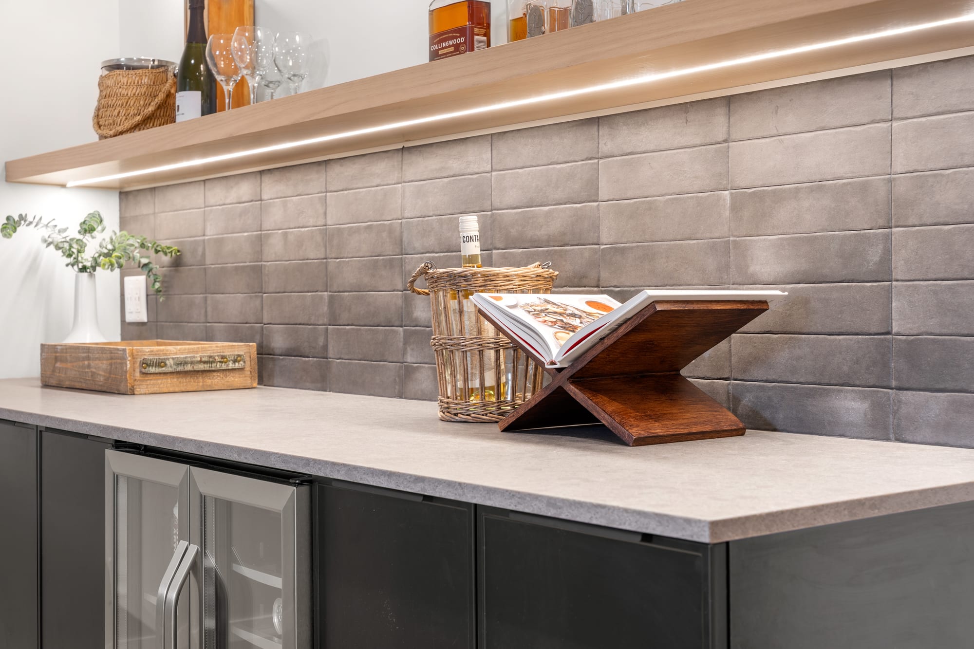 Wet bar detail with grey stacked tile backsplash and a lit floating oak shelf holding glassware and spirits