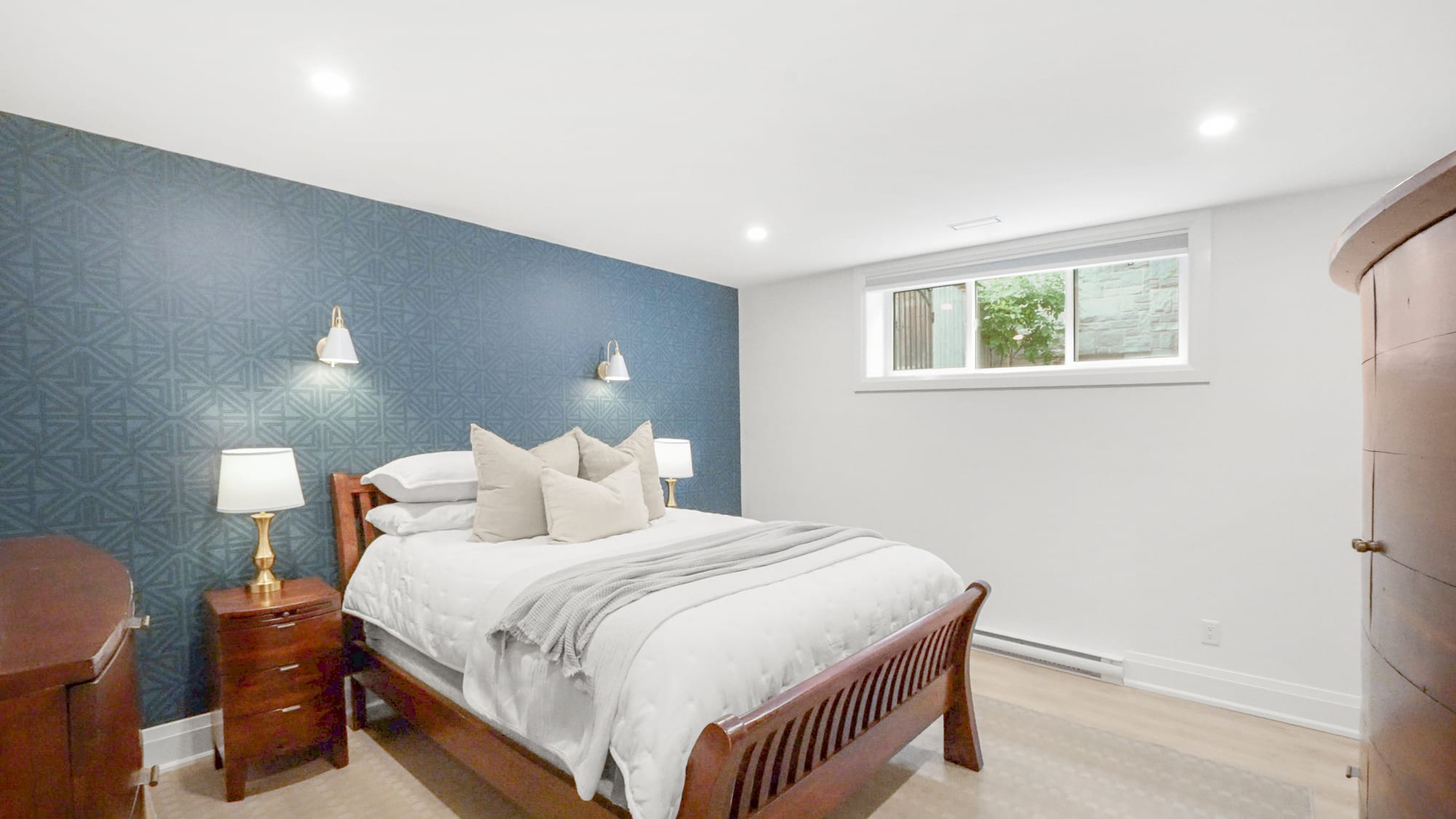 Lower-level bedroom with a patterned blue accent wall, brass wall sconces and an egress window