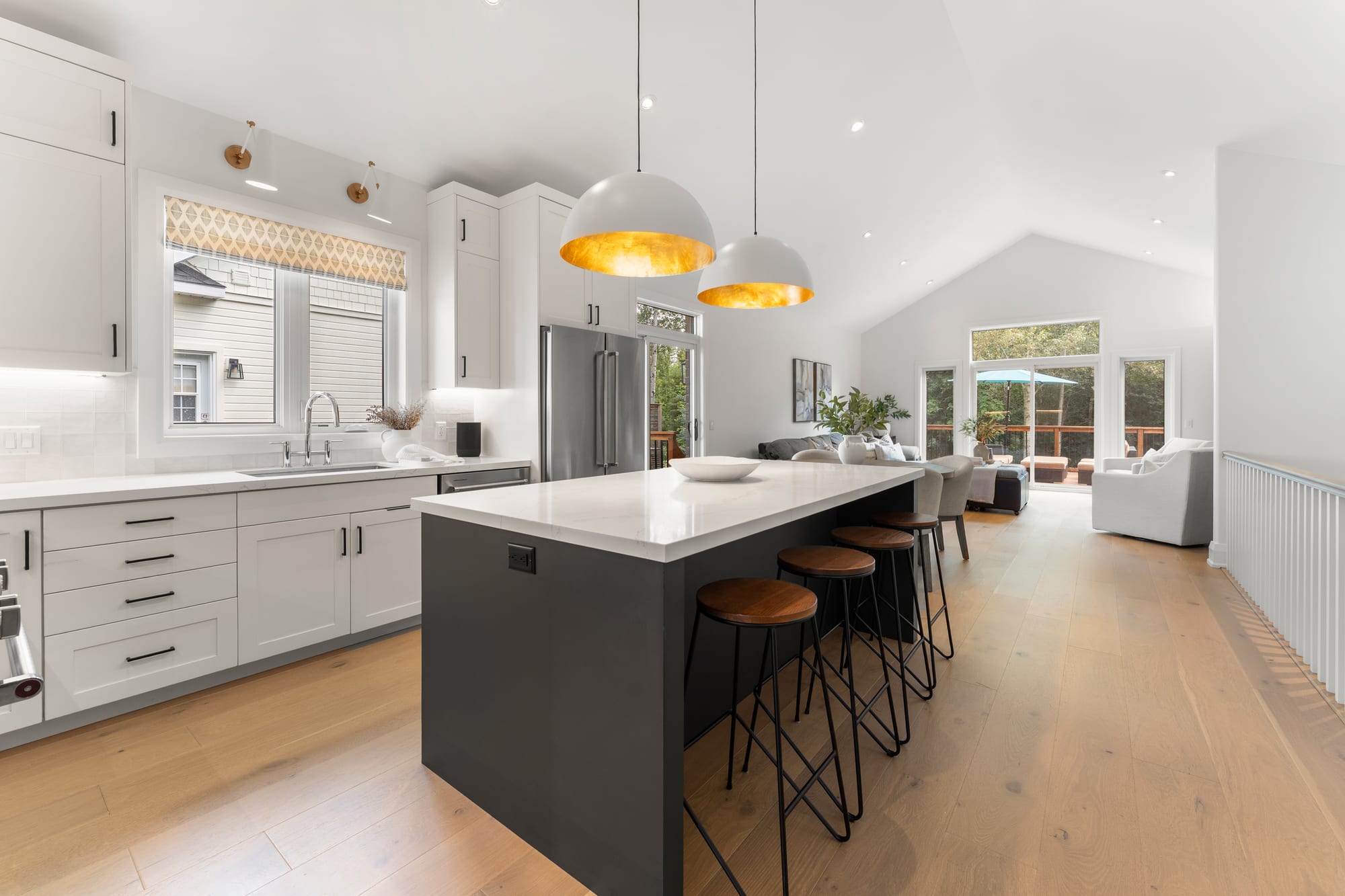 Kitchen sink under a window with quartz counters and shaker cabinetry, looking through to the vaulted great room and deck